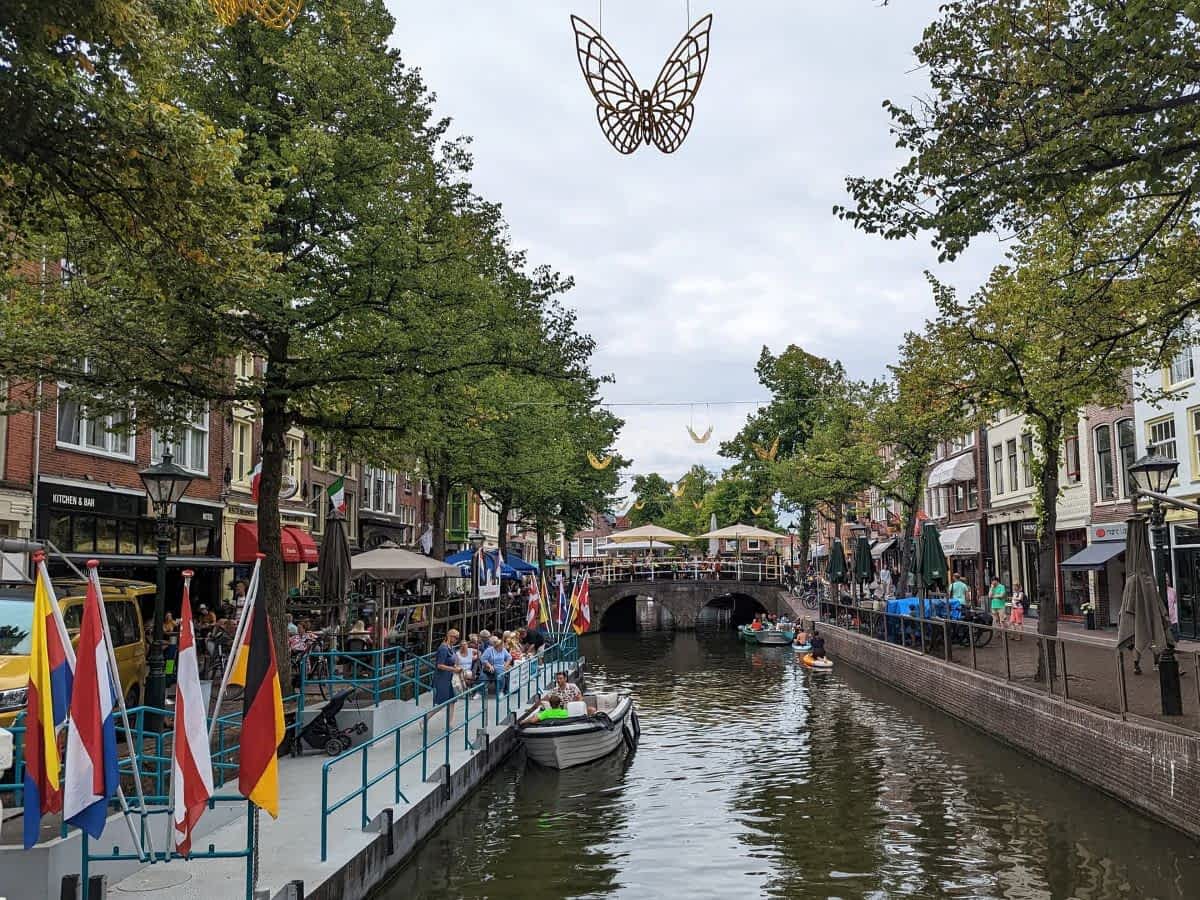 Blick auf eine belebte Gracht in Alkmaar mit Ausflugsbooten, schattigen Cafés am Ufer und dekorativen Schmetterlingen über dem Wasser.