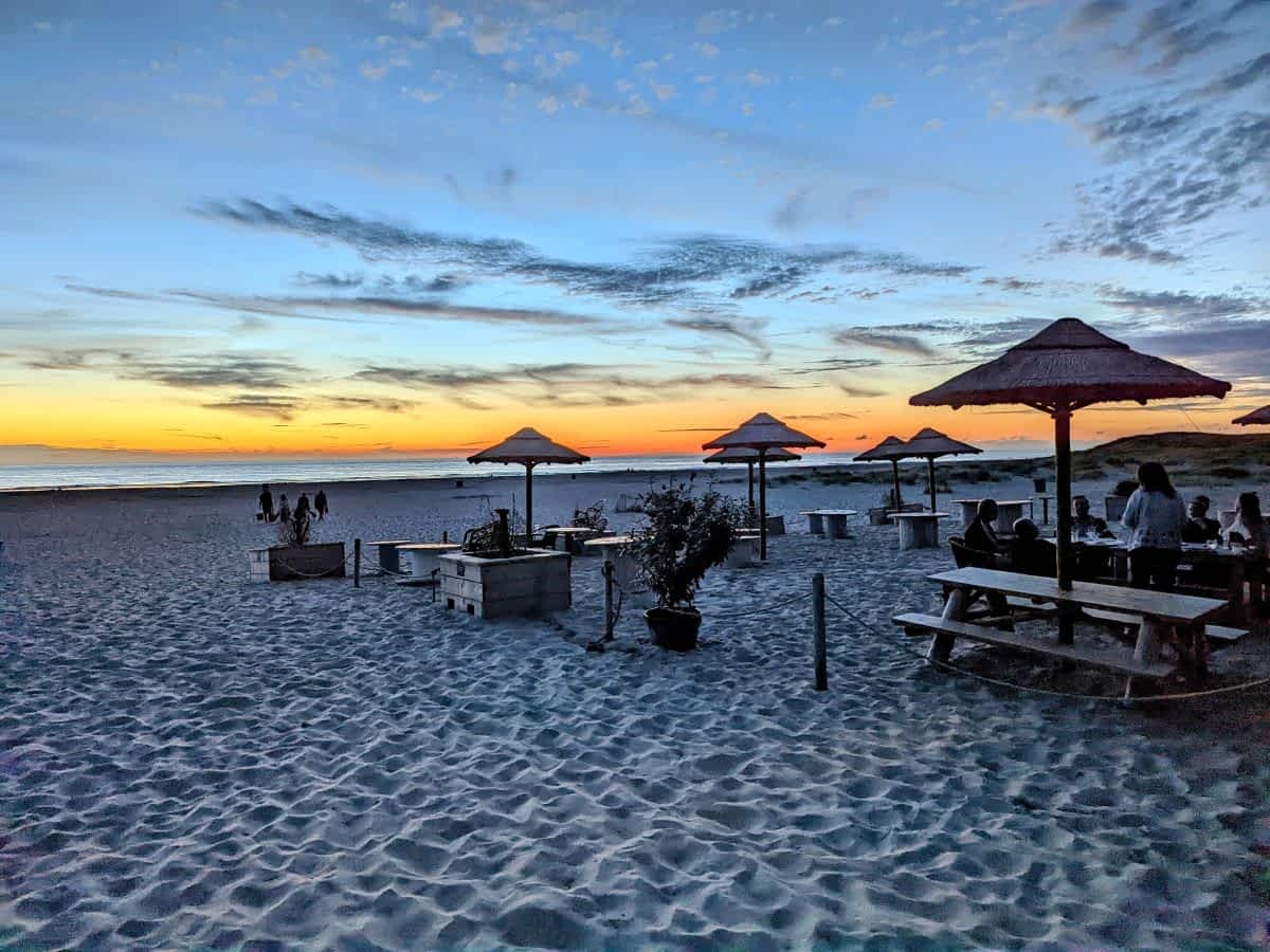 Strandbar in Hargen aan Zee bei Sonnenuntergang mit Holztischen, Bastschirmen und Blick auf die Nordsee.