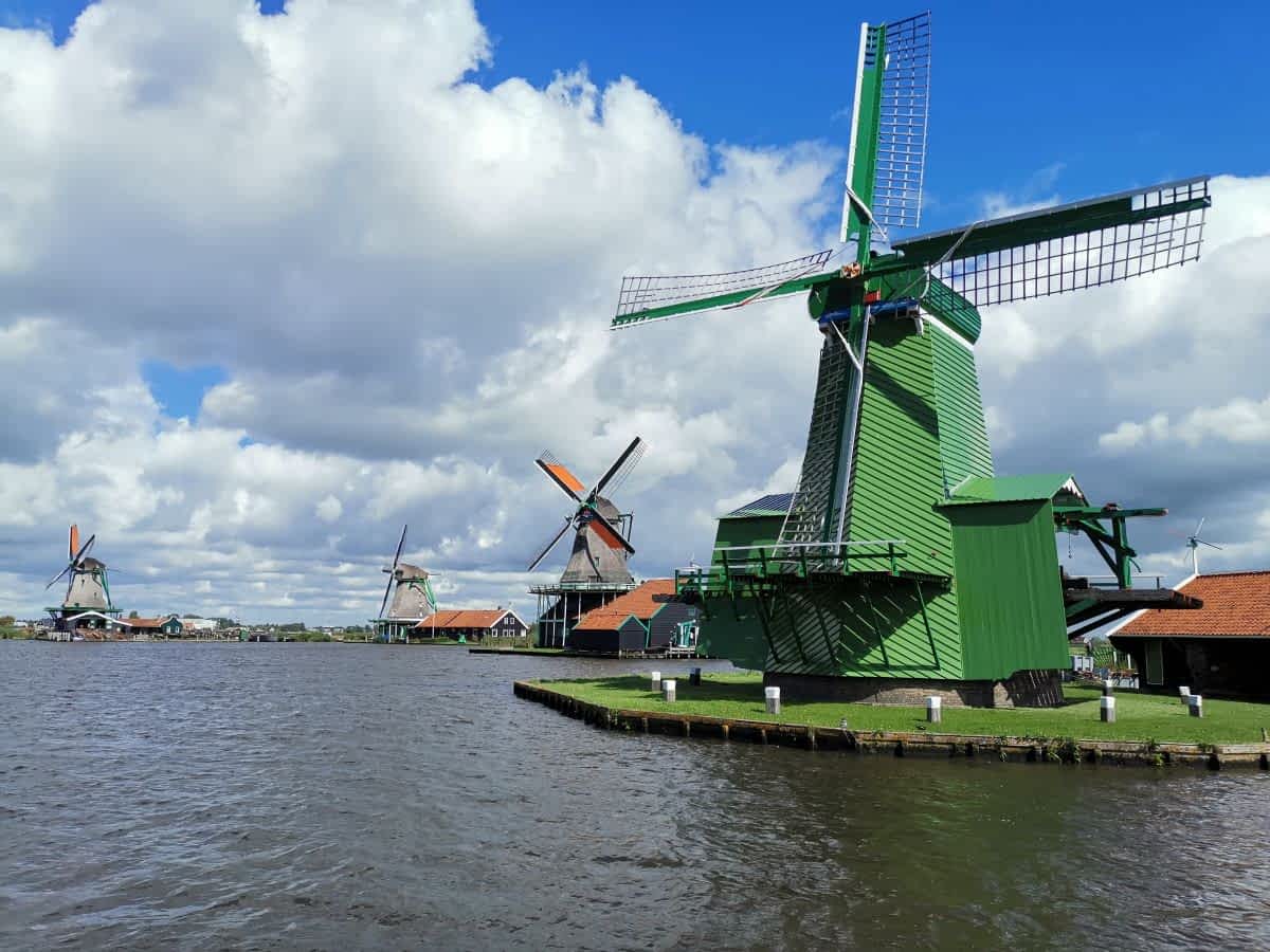 Blick über eine Gracht auf mehrere historische Windmühlen an der Zaanse Schans unter einem bewölkten Himmel. Im Vordergrund eine auffällig grüne Windmühle.