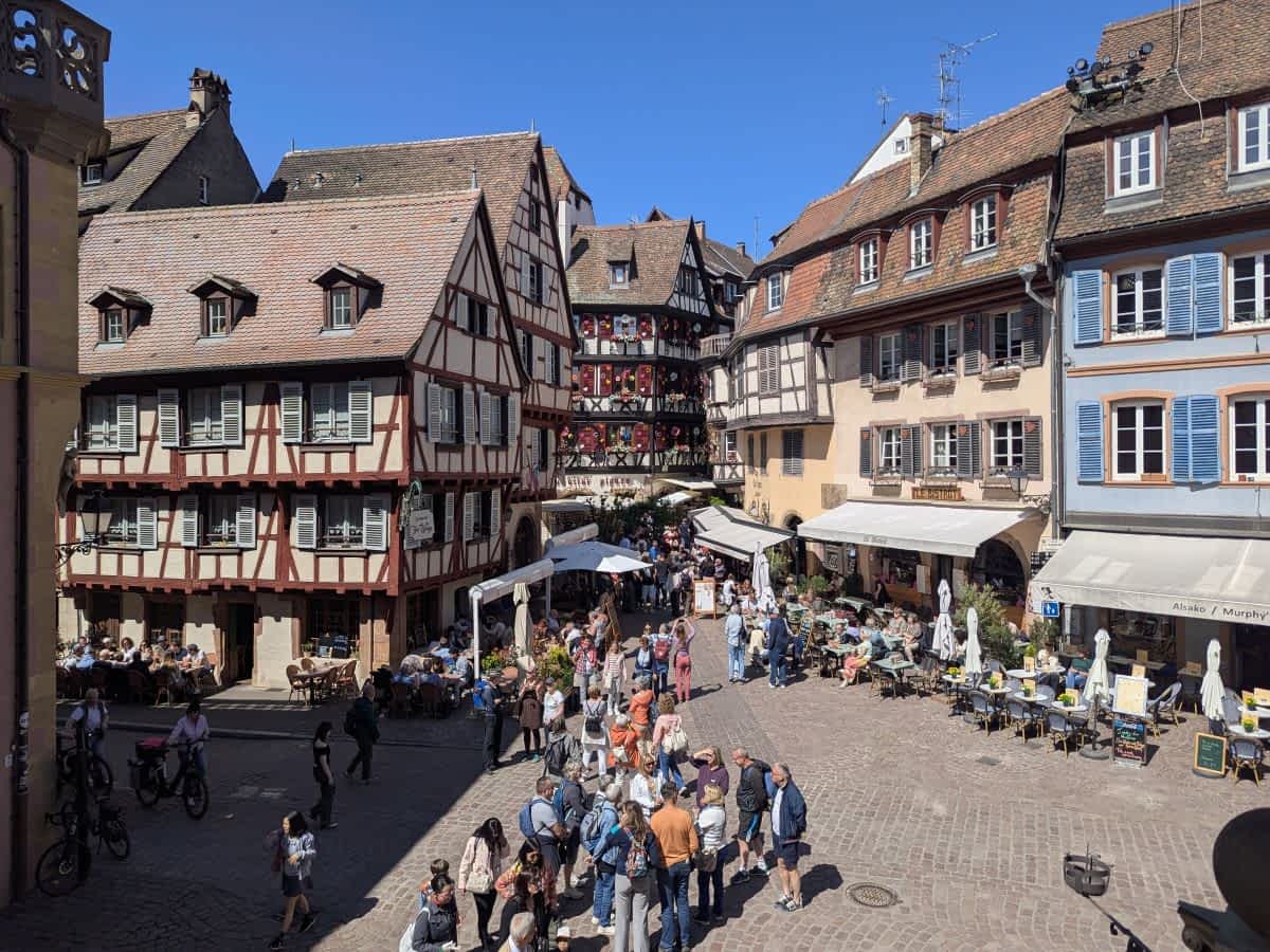 Belebter Platz in der Altstadt von Colmar mit zahlreichen Touristen, Cafés und prächtigen, blumengeschmückten Fachwerkhäusern unter blauem Himmel. Belebter Platz in der Altstadt von Colmar mit zahlreichen Touristen, Cafés und prächtigen, blumengeschmückten Fachwerkhäusern unter blauem Himmel.