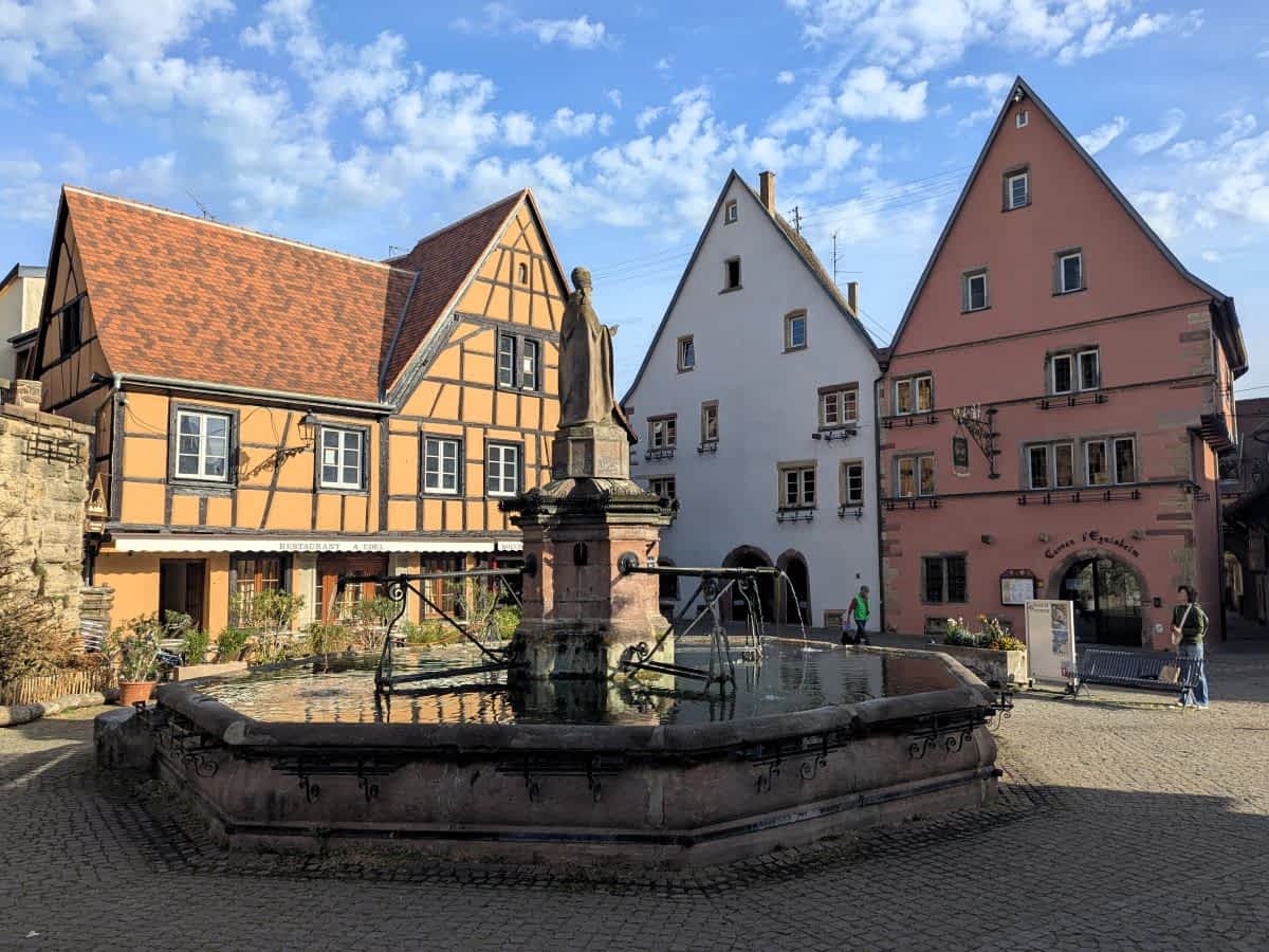 Der achteckige St.-Leo-Brunnen auf dem Marktplatz von Eguisheim vor farbenfrohen Fachwerkhäusern unter blauem Himmel. Der achteckige St.-Leo-Brunnen auf dem Marktplatz von Eguisheim vor farbenfrohen Fachwerkhäusern unter blauem Himmel.
