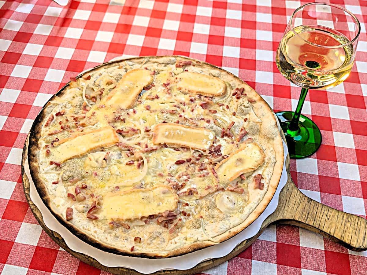 Ein elsässischer Flammkuchen mit Munsterkäse auf einem Holzbrett, daneben ein Glas Weißwein auf einer rot-weiß-karierten Tischdecke. Ein elsässischer Flammkuchen mit Munsterkäse auf einem Holzbrett, daneben ein Glas Weißwein auf einer rot-weiß-karierten Tischdecke.