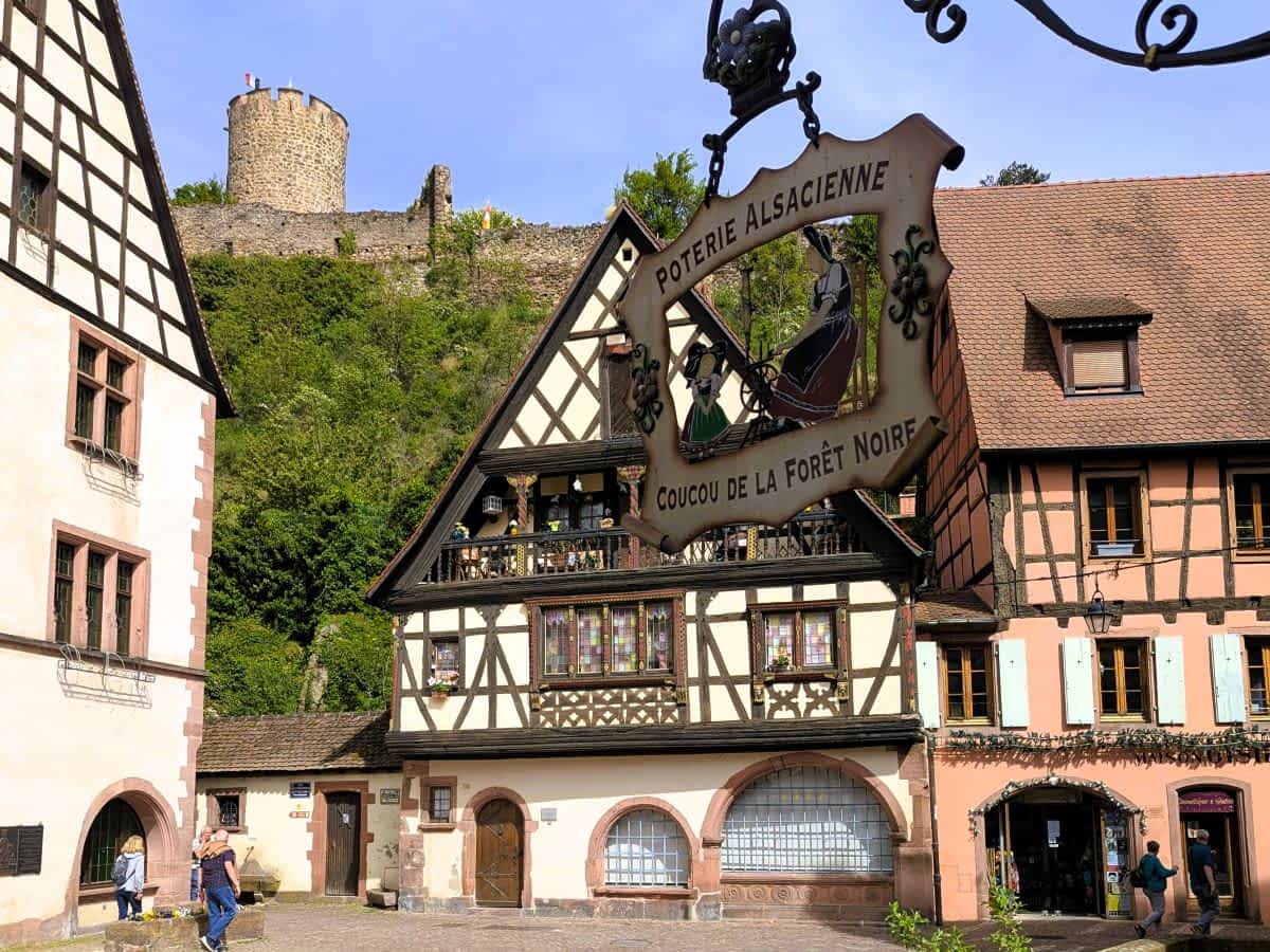 Historischer Platz in Kaysersberg mit prächtigen Fachwerkhäusern und der mittelalterlichen Burgruine auf dem bewaldeten Hügel im Hintergrund. Historischer Platz in Kaysersberg mit prächtigen Fachwerkhäusern und der mittelalterlichen Burgruine auf dem bewaldeten Hügel im Hintergrund.