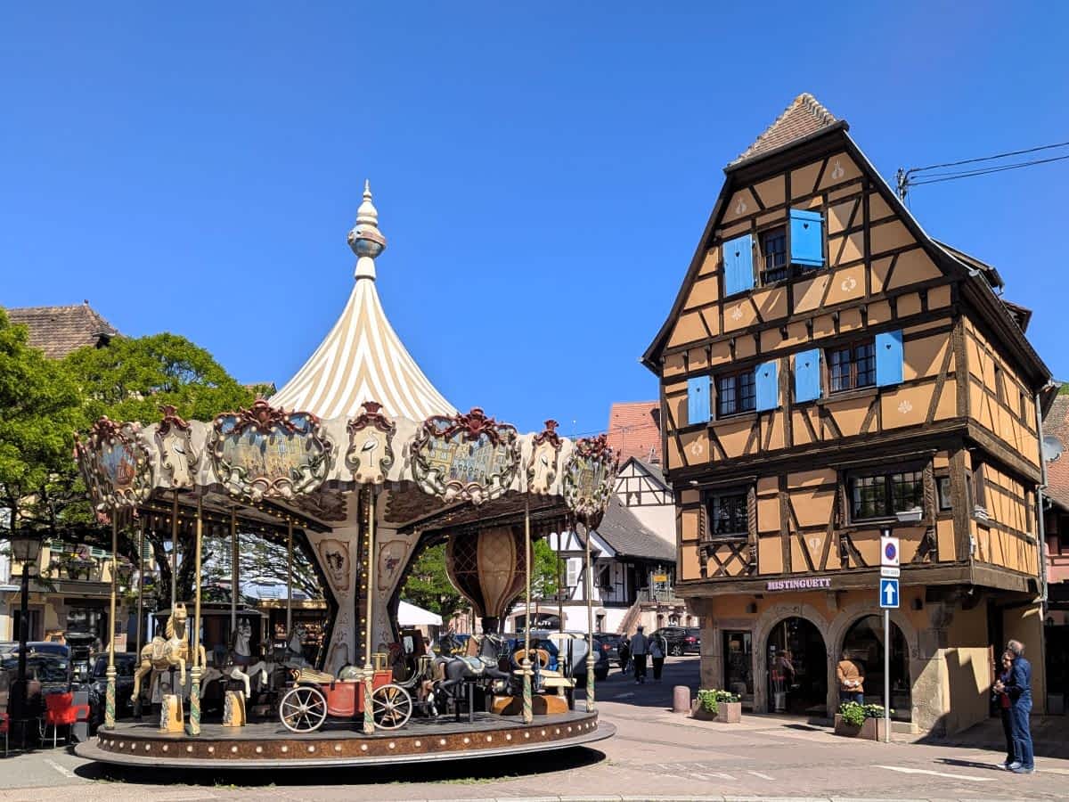Ein nostalgisches, weiß-gestreiftes Karussell auf einem öffentlichen Platz in Obernai neben einem hohen Fachwerkhaus mit blauen Fensterläden. Ein nostalgisches, weiß-gestreiftes Karussell auf einem öffentlichen Platz in Obernai neben einem hohen Fachwerkhaus mit blauen Fensterläden.