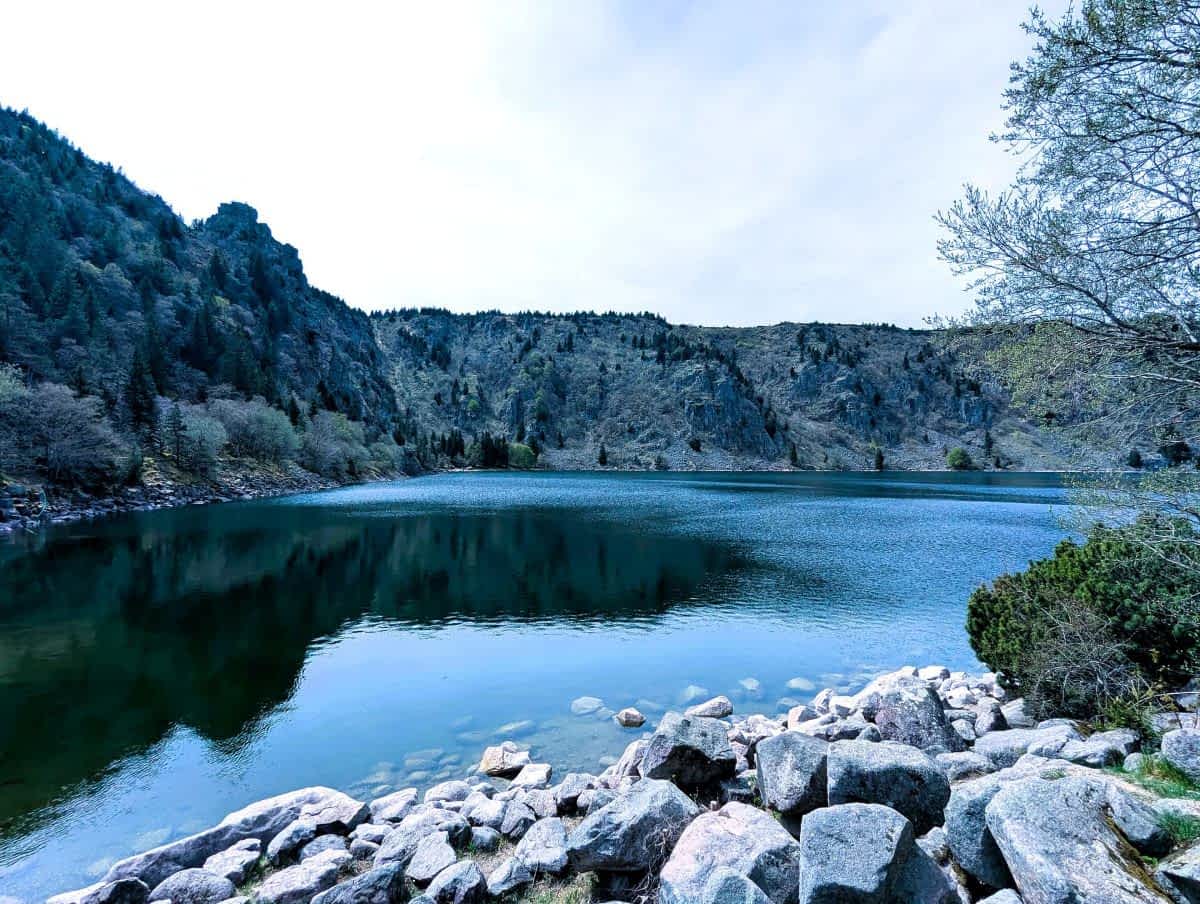 Ein klarer, blauer Bergsee (Lac Blanc) in den Vogesen, umgeben von steilen, bewaldeten Felswänden und grauen Granitblöcken im Vordergrund Ein klarer, blauer Bergsee (Lac Blanc) in den Vogesen, umgeben von steilen, bewaldeten Felswänden und grauen Granitblöcken im Vordergrund